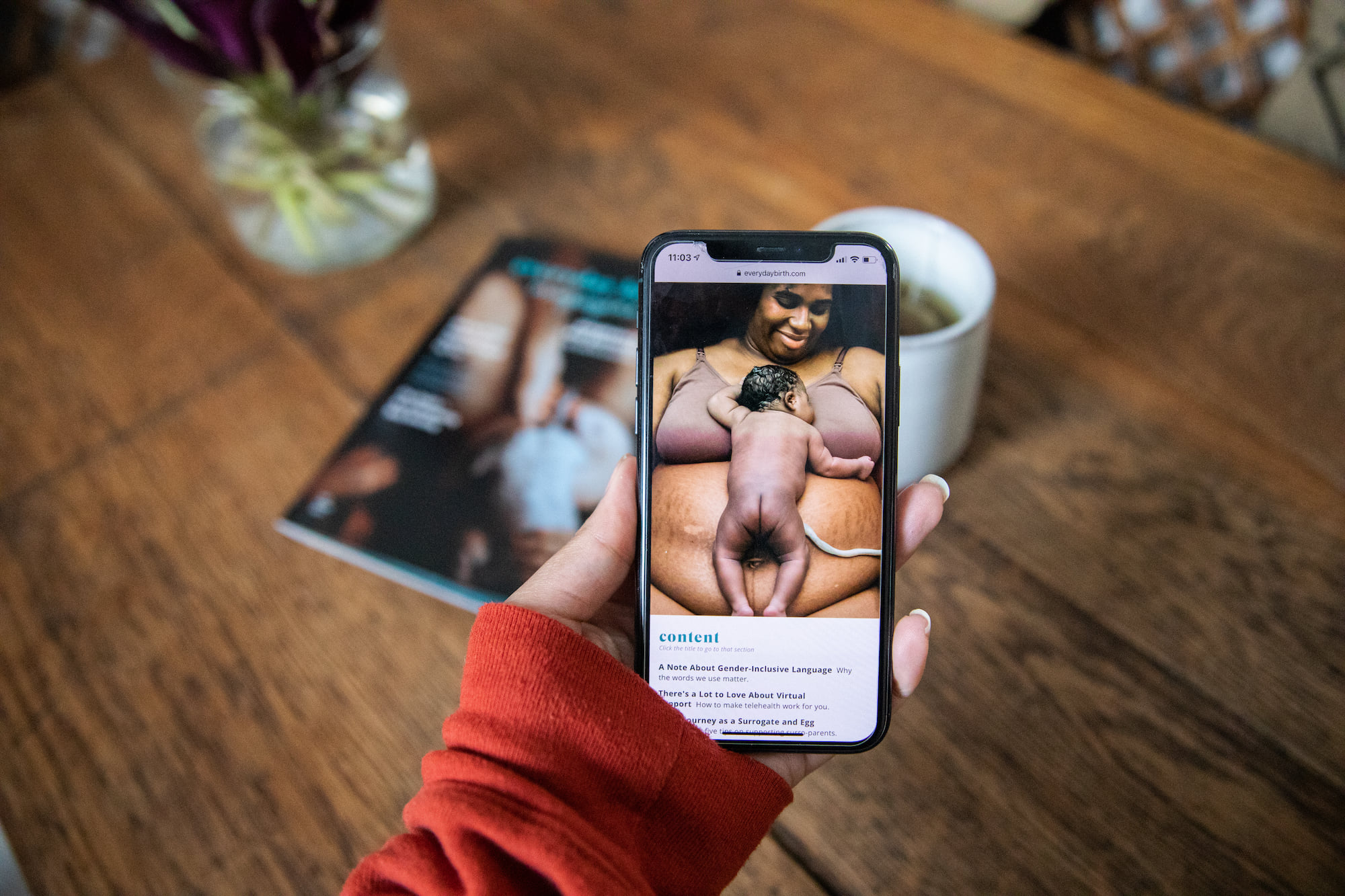 Hand holding a phone with an image from the magazine — a new plus-size parent happily holding their newborn on their chest.