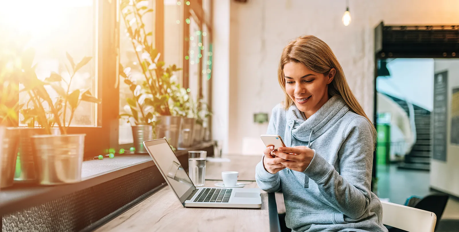 Woman smiling, looking at her phone with her laptop open on the table next to her.